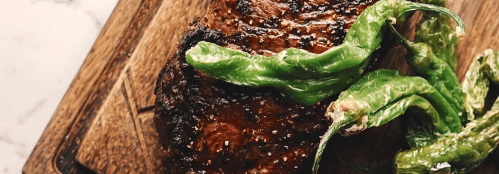 Savage Chef Grilled steak with charred shishito peppers on a wooden board.