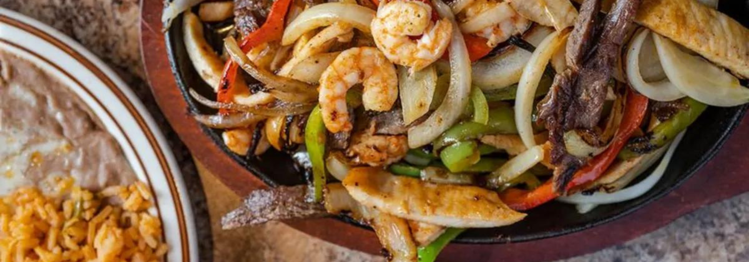 Untitled design (91) A sizzling platter of mixed fajitas with beef, chicken, shrimp, onions, and bell peppers, served alongside a plate of refried beans and rice.