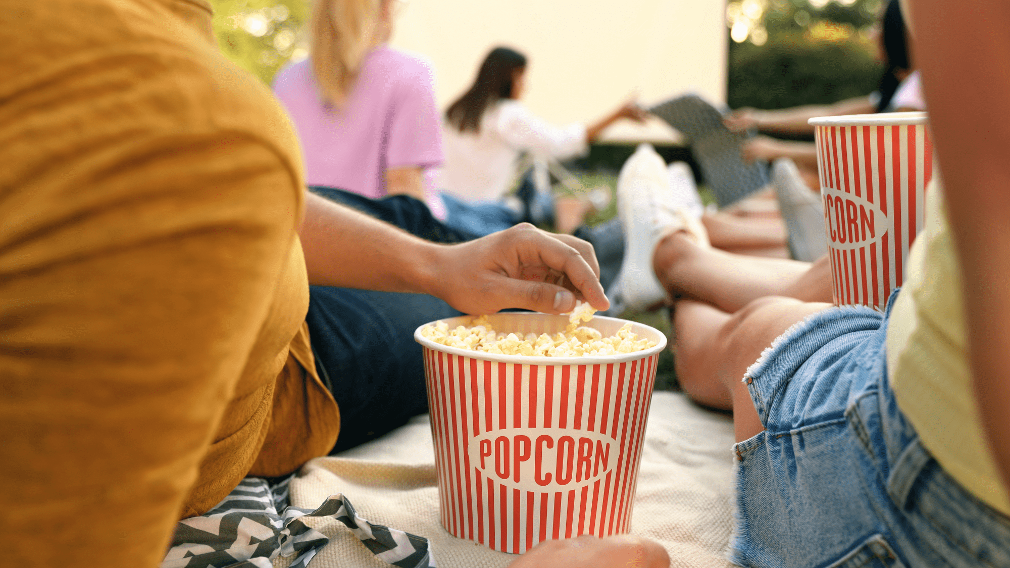 People lounging on a blanket outside, sharing popcorn from striped containers, with a screen set up in the background.